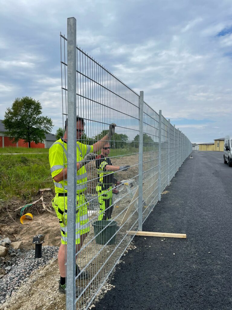 Två män i arbetskläder som monterar ett stängsel i metall.
